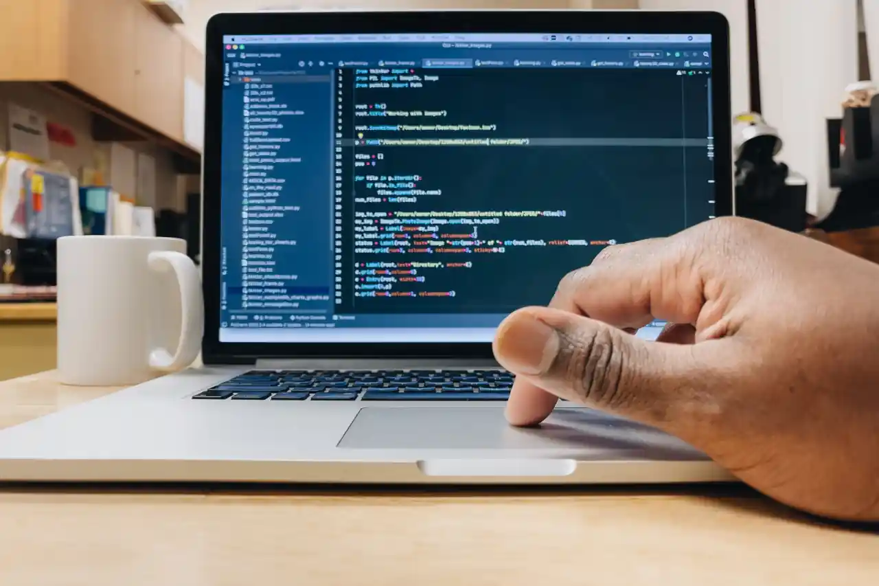 Hand am Trackpad eines Laptops, auf dem ein Code-Editor geöffnet ist